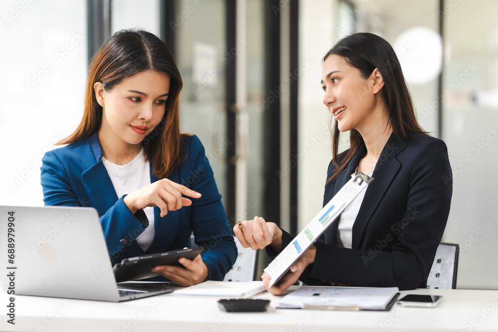 Two middle aged and young Asian female executives in formal suits review bar chart, discussing business strategies in office setting, senior executives or directors in advertising or public relations Two middle aged and young Asian female executives in formal suits review bar chart, discussing business strategies in office setting, senior executives or directors in advertising or public relations