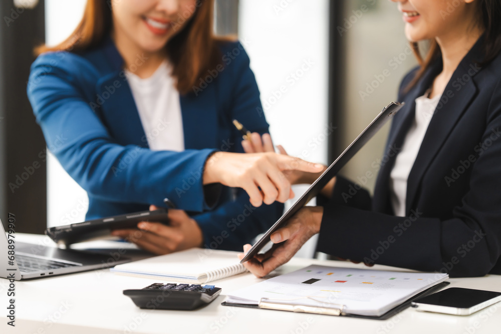 Fototapeta premium Two middle aged and young Asian female executives in formal suits review bar chart, discussing business strategies in office setting, senior executives or directors in advertising or public relations
