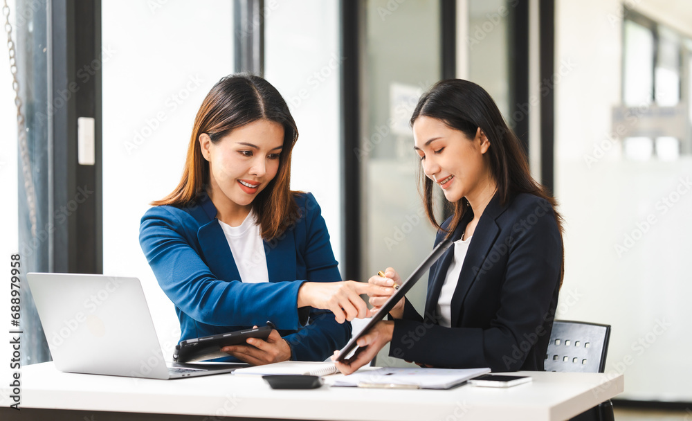 Two middle aged and young Asian female executives in formal suits review bar chart, discussing business strategies in office setting, senior executives or directors in advertising or public relations Two middle aged and young Asian female executives in formal suits review bar chart, discussing business strategies in office setting, senior executives or directors in advertising or public relations