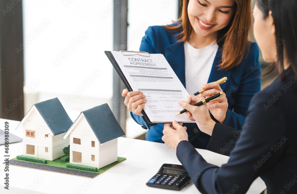 Two Asian real estate agents in formal suits discuss a contract with house models on the table, ready to sign a deal. Two Asian real estate agents in formal suits discuss a contract with house models on the table, ready to sign a deal.