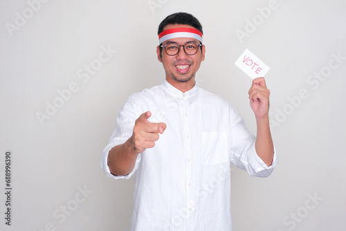 Indonesian man pointing at the camera while holding election vote card with happy expression photo