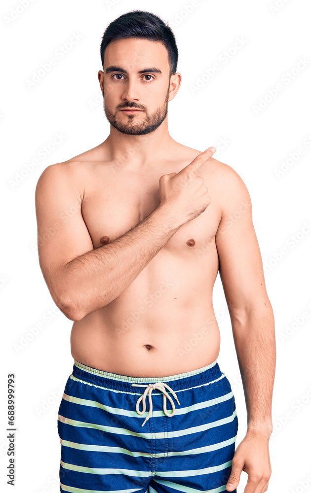 Young handsome man wearing swimwear pointing with hand finger to the side showing advertisement, serious and calm face Young handsome man wearing swimwear pointing with hand finger to the side showing advertisement, serious and calm face