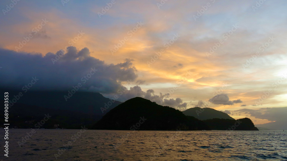 Sunset over the mountains. Sunset over the sea and tropical island. The sun behind the clouds sets below the horizon. The sun's rays color the sky and clouds during sunset.