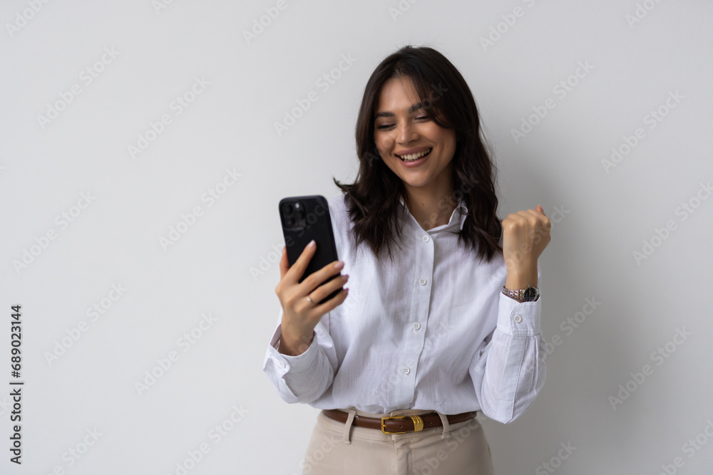 Enthusiastic woman winning on phone rejoicing and screaming of joy while using phone app, standing over white background Enthusiastic woman winning on phone rejoicing and screaming of joy while using phone app, standing over white background