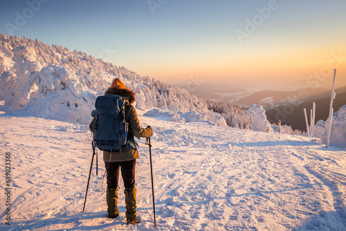 Woman with backpack and nordic walking poles trekking in cold weather. Hiking in snow at winter mountain during sunset. Sports and outdoors seasonal activity