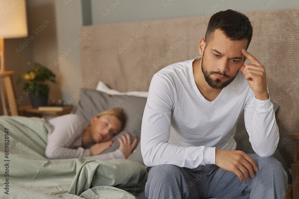 Young man deep in thoughts while his wife sleeps in the background Young man deep in thoughts while his wife sleeps in the background
