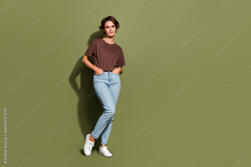 Full size photo of pretty woman dressed brown t-shirt arms in pockets stand near offer empty space isolated on khaki color background Full size photo of pretty woman dressed brown t-shirt arms in pockets stand near offer empty space isolated on khaki color background