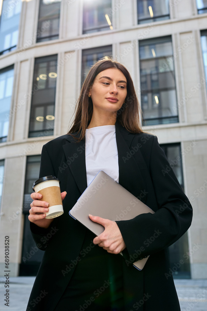 A successful business woman takes a moment to recharge, standing with a laptop and a coffee cup outside a busy office complex A successful business woman takes a moment to recharge, standing with a laptop and a coffee cup outside a busy office complex