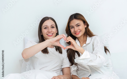 Young Asian lesbian couple sitting on bed during happy resting time, hand made heart symbol. Be happy and relaxed in the bedroom.