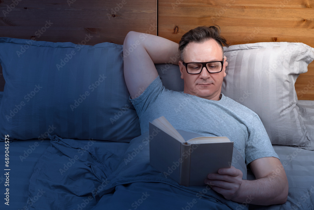 Fototapeta premium A man with glasses is lying on the bed reading a book before going to bed, by the light of a night light