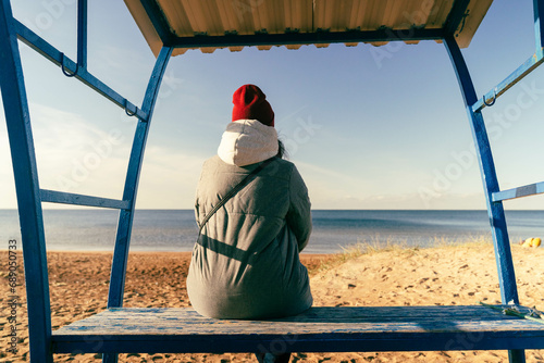 Fototapeta Naklejka Na Ścianę i Meble -  woman in an autumn jacket and hat is sitting alone on a bench on a deserted autumn beach on a sunny day