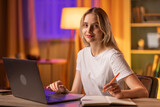 A pretty smiling blonde girl student sits at a table in apartment, uses a laptop to search textbooks and teaching aids. A young lady blogger communicates with subscribers and take notes suggestions