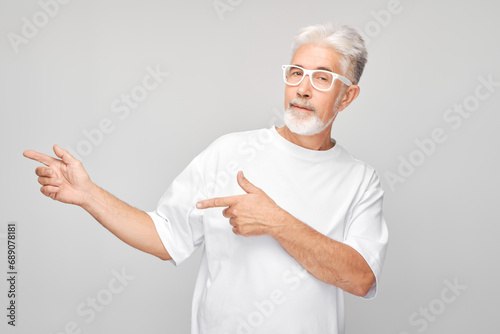 Gray-haired mature man 60 years old in white T-shirt pointing aside finger, demonstrating empty space for product or text isolated on white studio background. © amixstudio