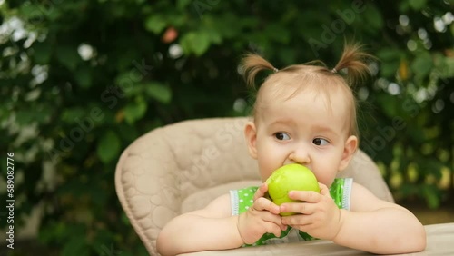 Portrait of a cute funny baby girl, eating a green apple, sitting in a high chair in the garden on a sunny summer day.