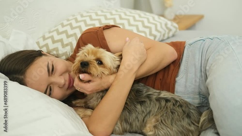 A young smiling woman hugs a Yorkshire terrier lying at home on a bed.