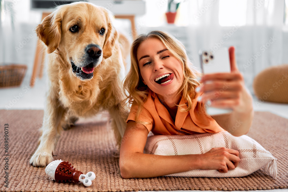 Fun together. A happy woman is taking a selfie with her cheerful pet, who has a funny expression on his face, and they are lying on the living room floor and making funny faces.Emotional connection. - obrazy, fototapety, plakaty 