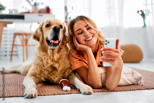Saving memories with pet. Smiling woman with blond hair snuggling to furry friend and taking selfie on modern cell phone. Obedient golden retriever lying on floor near delighted female owner.