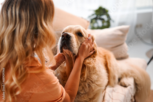 Shared moments. Back view of female blonde caressing furry dog behind ears during leisure time at cozy apartment. Young woman and golden retriever enjoying bonding interaction together during daytime. #689091966