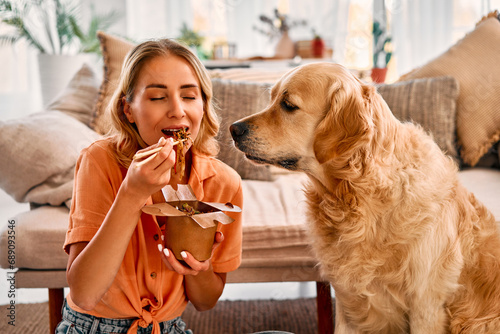 Pet and owner at home. Pleasant young woman with wavy blond hair sitting on floor and eating chinese noodles at living room. Adult golden retriever with begging eyes sniffing aroma human food. photo