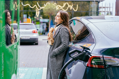 Slim brunette girl eating fast food hotdog or burger while waiting electric vehicle to charge. outdoor on the street.