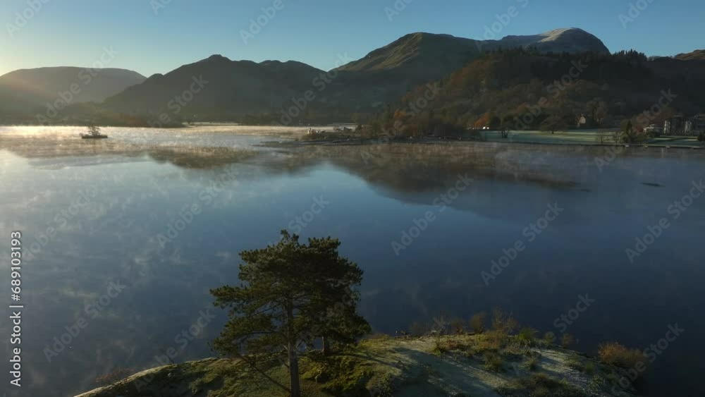 Flyover of small island in calm lake with mist gently curling on water surface at sunrise in autumn. Ullswater, English Lake District, Cumbria, UK. Flyover of small island in calm lake with mist gently curling on water surface at sunrise in autumn. Ullswater, English Lake District, Cumbria, UK.