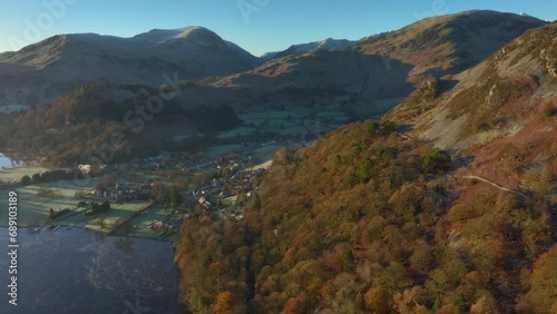 Autumn sunrise sunshine on hillside trees with lakeside town backed by mountains. Glenridding, Ullswater, English Lake District, Cumbria, UK. photo