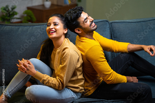 Laughing indian couple talking each other by sitting back to back on sofa at home during evening - concept of happiness, dating and relationship bonding