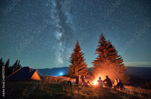 Night camping in mountains under starry sky. Back view of group of people traveling. Friends sitting in campsite near fire  talking  enjoying nature. Concept of tourism  adventure and traveling.