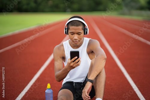 Asian man sportsman fit body wearing white headphone and using mobile phone on white screen to listen music while resting or before jogging, exercising at lane stadium. Sport healthy running concept. photo