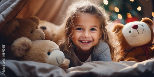 A charming cheerful little girl dressed in pajamas, playing with soft toys and bears on the bed, playing hide-and-seek, hiding under the blanket. The child is having fun at home. The concept of holida