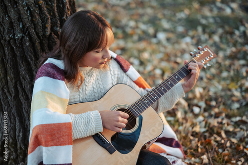 Young girl playing the guitar near the tree in autumn park. Beautiful evening shot of a girl musician outdoors in autumn nature