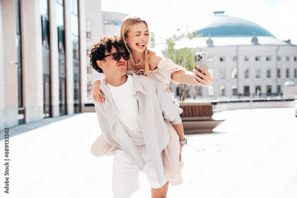 Fototapeta premium Young smiling beautiful woman and her handsome boyfriend in casual summer clothes. Happy cheerful family. Couple posing in street at sunny day. Female sits at his back. piggyback riding. Take selfie
