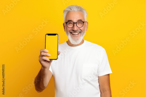 Happy older senior business man wearing shirt holding smartphone pointing at mock up screen mobile display on yellow background
