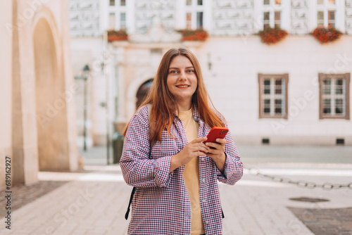 Picture of pretty young woman staying on the street holding phone in hands. 30s tourist walking on old city street checks her smartphone. Use technology concept, Traveling Europe in summer