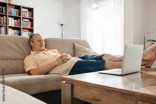 A man is relaxing after a working day, sitting on the couch and watching a TV show or movie through a laptop.