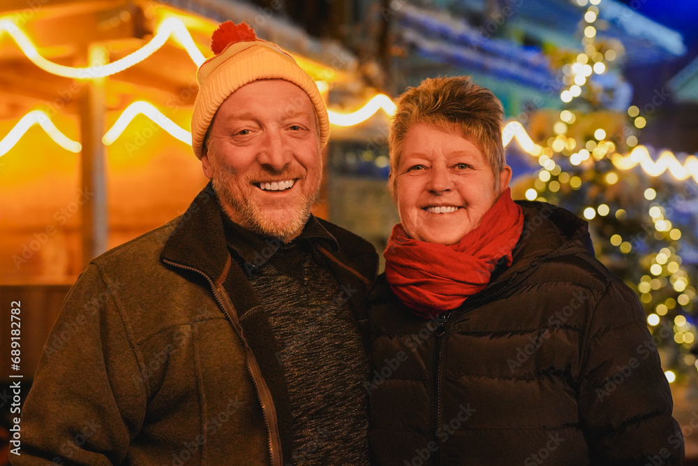 Happy senior couple smiling on camera during winter night time outside with christmas tree and lights in the background