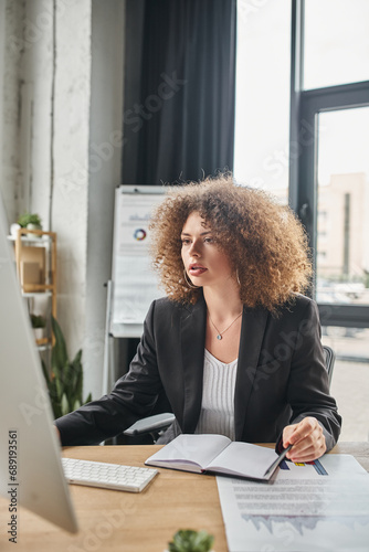 young ambitious businesswoman with wavy hair working on computer at workplace in modern office