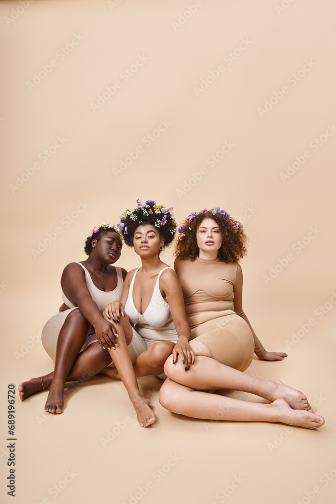 body positive multiethnic girlfriends in lingerie with flowers in hair sitting on beige backdrop