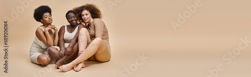 full length of multicultural plus size girlfriends in underwear sitting on beige, horizontal banner