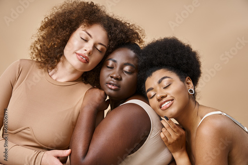 multiracial body positive women in lingerie smiling with closed eyes on beige, plus size beauty
