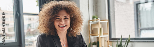 portrait of joyful businesswoman with curly hair looking at camera in modern office, banner