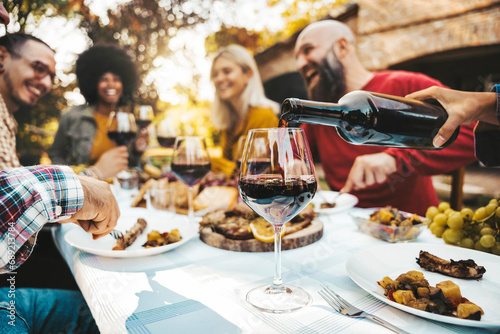 Happy friends having bbq dinner party in garden restaurant - Cheerful family drinking red wine together sitting at dining table - Food and beverage concept with guys and girls eating fresh meal photo