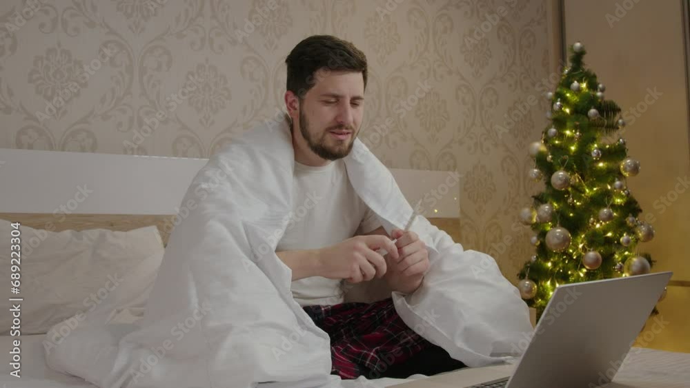 Sad and cold man in pajamas at home sprays spary in his nose. Sick boy with fever is wrapped, watches a movie on the computer and celebrates the winter holidays at home in a calm atmosphere