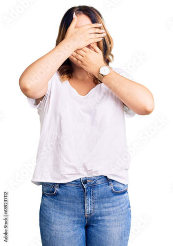 Young beautiful caucasian woman wearing casual white tshirt covering eyes and mouth with hands, surprised and shocked. hiding emotion