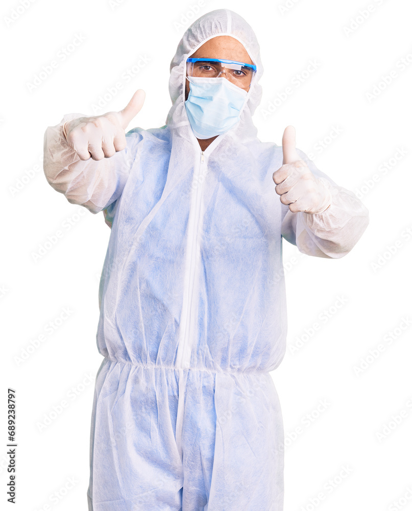 Young hispanic man wearing doctor protection coronavirus uniform and medical mask approving doing positive gesture with hand, thumbs up smiling and happy for success. winner gesture. Young hispanic man wearing doctor protection coronavirus uniform and medical mask approving doing positive gesture with hand, thumbs up smiling and happy for success. winner gesture.