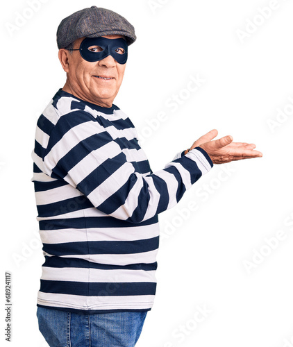 Senior handsome man wearing burglar mask and t-shirt pointing aside with hands open palms showing copy space, presenting advertisement smiling excited happy © Krakenimages.com