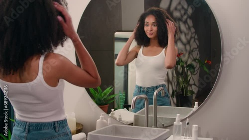 African American woman come in home bathroom looking at mirror reflection beautiful pretty ethnic girl female lady fix curls check curly hair doing hairdo hairstyle beauty routine shampoo go away photo