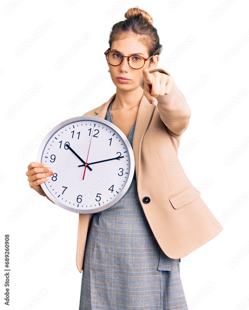Beautiful caucasian woman with blonde hair wearing business jacket and holding clock pointing with finger to the camera and to you, confident gesture looking serious Beautiful caucasian woman with blonde hair wearing business jacket and holding clock pointing with finger to the camera and to you, confident gesture looking serious