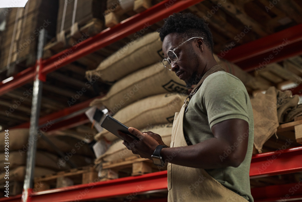 Side view portrait of young African American man holding digital tablet at warehouse in coffee roastery, copy space Side view portrait of young African American man holding digital tablet at warehouse in coffee roastery, copy space