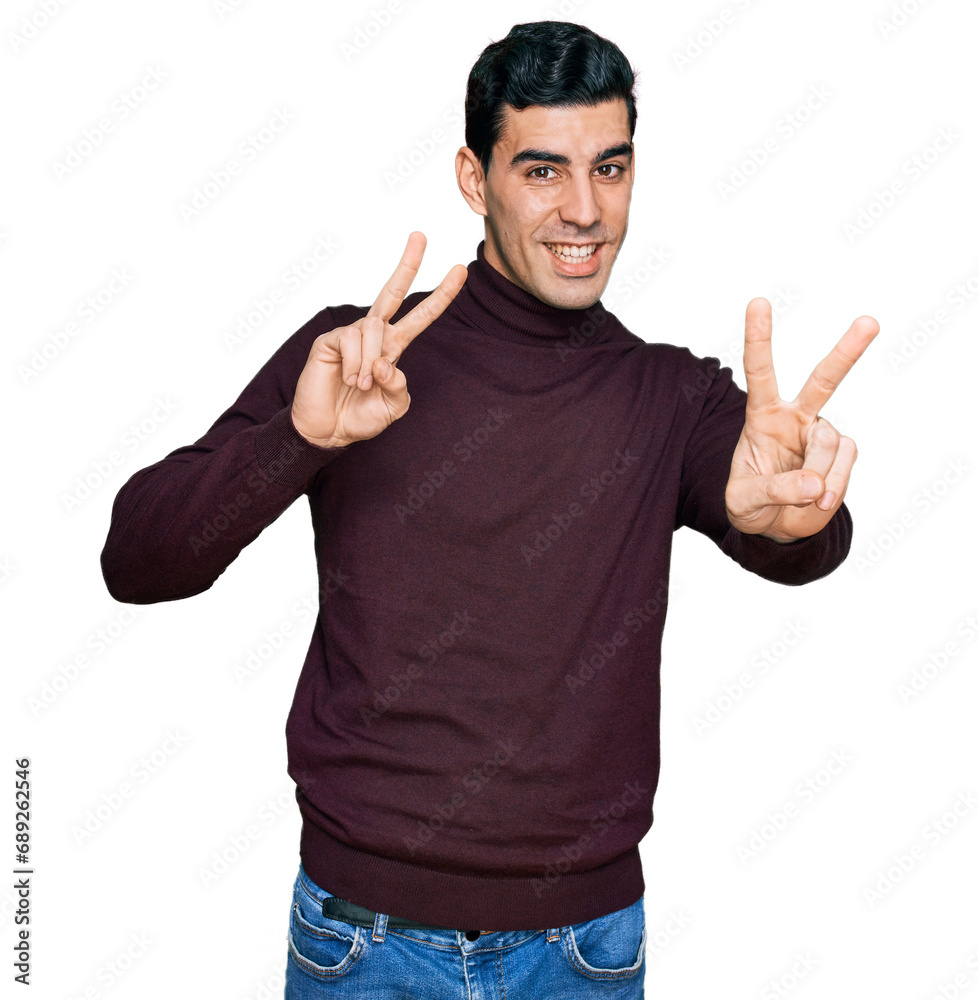 Fototapeta premium Handsome hispanic man wearing casual turtleneck sweater smiling looking to the camera showing fingers doing victory sign. number two.